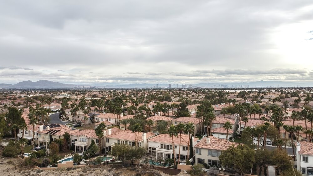 Aerial view of a suburban area with numerous houses featuring red-tiled roofs, surrounded by palm trees and greenery. The background, resting under a cloudy sky, hints at a distant city skyline—an environment made peaceful through reliable pest control.