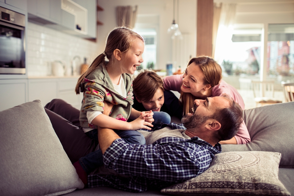 A family of four enjoys a playful moment on a couch in their modern living room, free from worries thanks to reliable pest control. The father lies back while the mother, son, and daughter laugh together in the bright, airy space with a kitchen visible in the background.