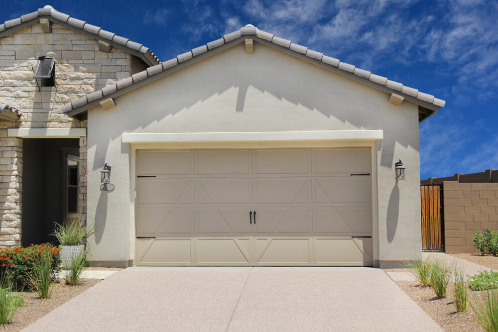 A modern house with a light-colored exterior and a two-car garage offers an inviting look. The roof is tiled, with reliable wall lanterns flanking the garage door. The concrete driveway, framed by landscaped areas, adds charm to this residence that embodies effective solutions for contemporary living.