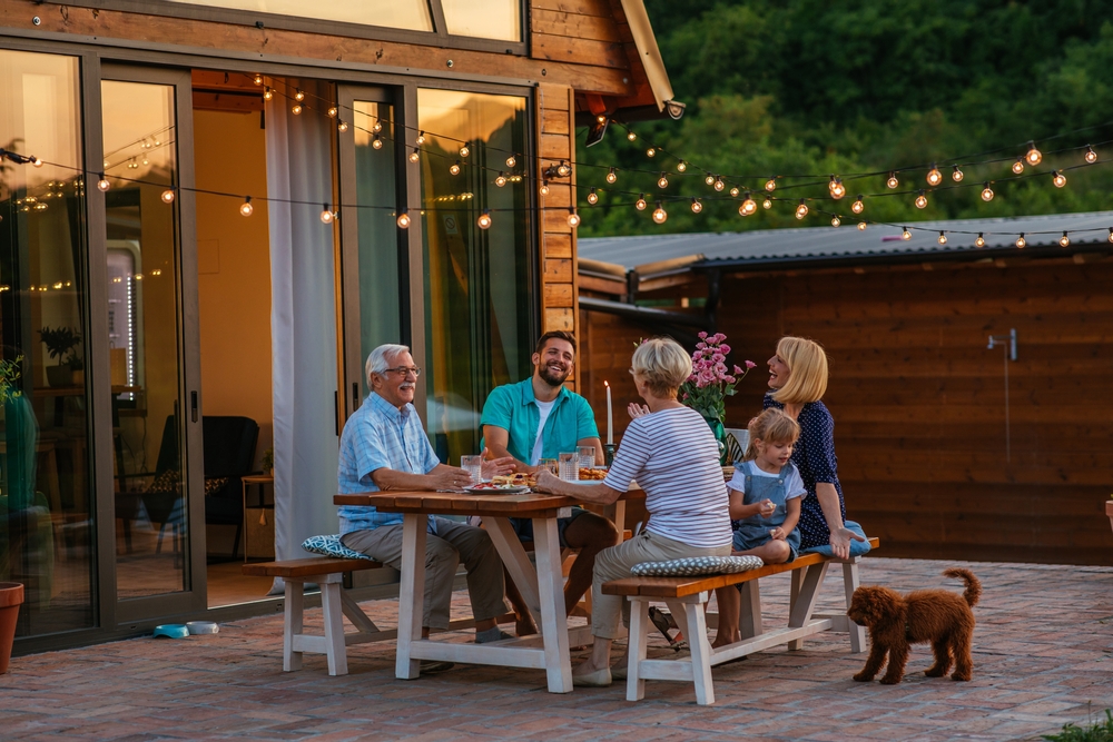 A group of people sit around a picnic table on a patio, enjoying a meal free from mosquito nuisances. String lights hang above them, and a potted plant graces the table. A child is present, as a small brown dog approaches. In the background, a wooden building stands surrounded by trees.